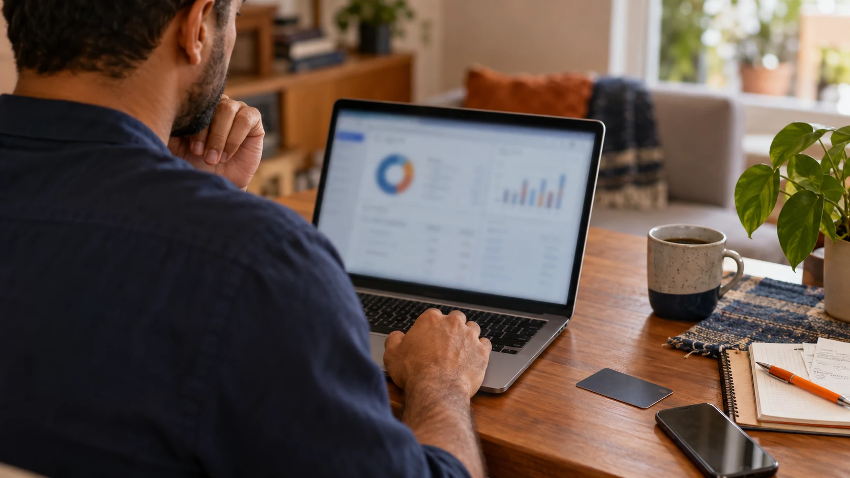 Homem consultando informações financeiras no computador, com documento virado sobre a mesa.